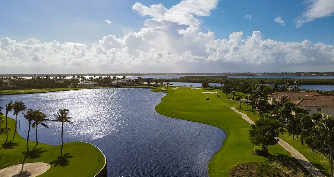 Elevated view of a golf fairway curving around a water hazard with palm trees, bunkers, and houses.