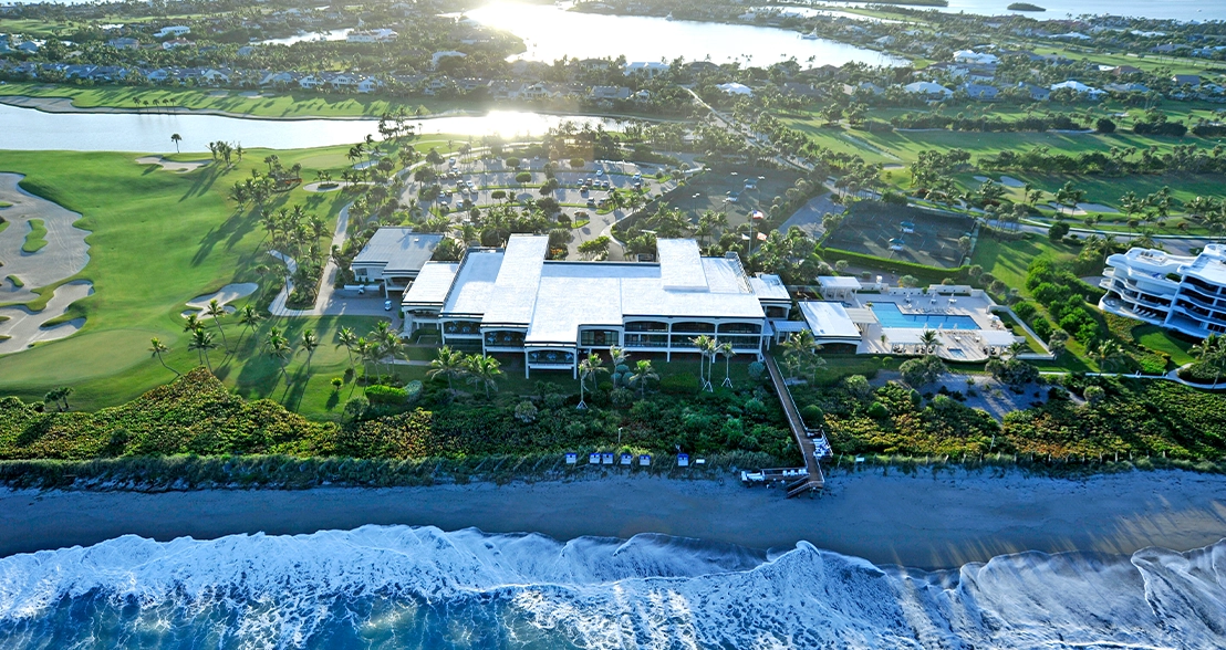 Aerial view of an oceanfront clubhouse with beach waves, golf course, palm trees, and inland lagoons.