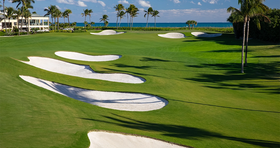 Oceanfront golf green with white sand bunkers, palm trees, and a blue sea horizon.