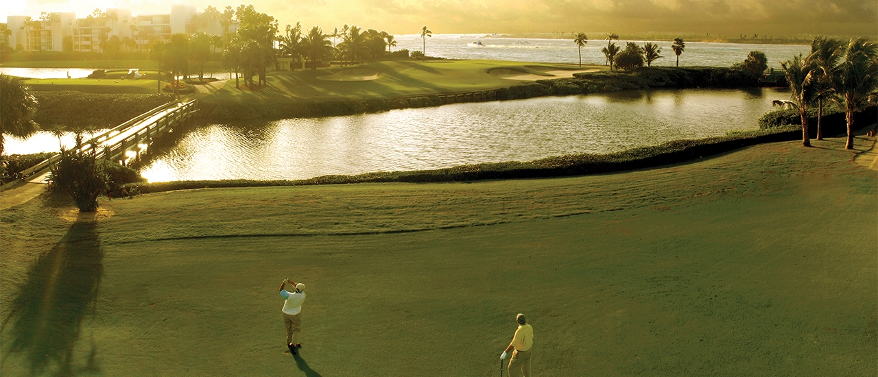 Two golfers stand on a fairway near a pond at sunset, with one golfer mid-swing.